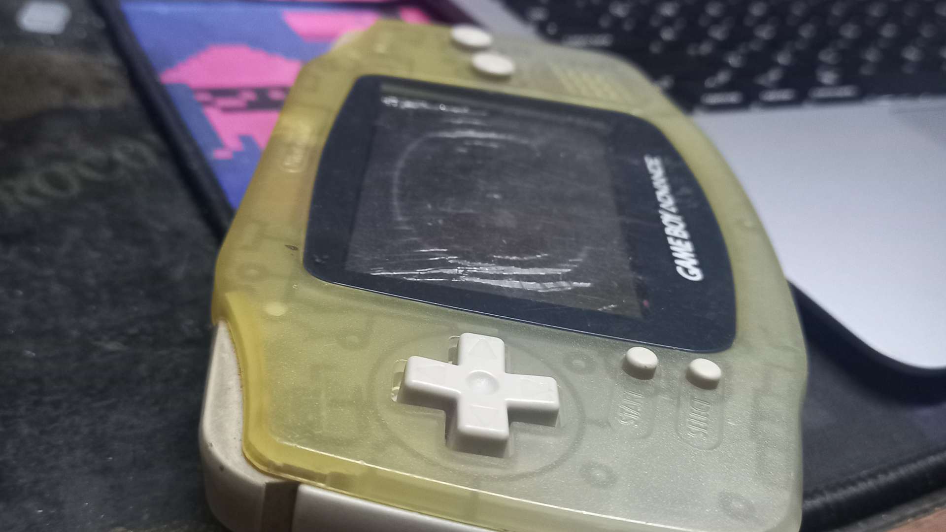 A worn translucent yellow-green Game Boy Advance with a cracked screen, resting on a desk beside a keyboard